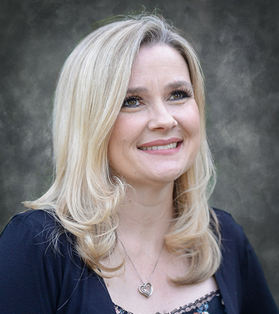 A woman with long, straight blonde hair and light makeup is smiling slightly while looking upward and slightly to her right. She is wearing a dark top with a floral pattern and a silver heart pendant necklace.