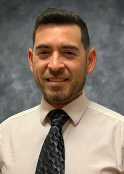 Man with short cropped hair and five o clock shadow wearing a white shirt with solid, dark tie